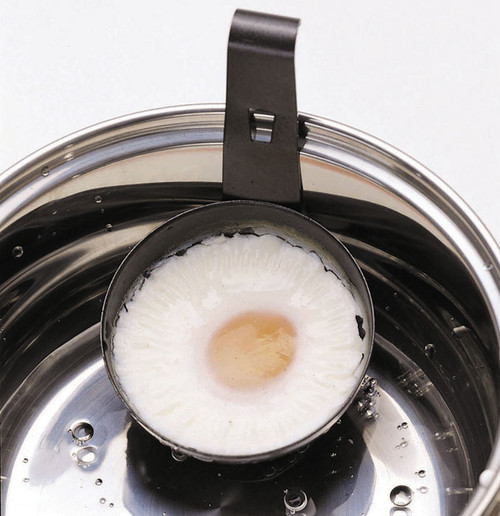 Black non-stick egg poacher cup with a metal hanging hook, holding a cooked poached egg with a pale yellow yolk, positioned inside a stainless steel saucepan with water droplets visible.