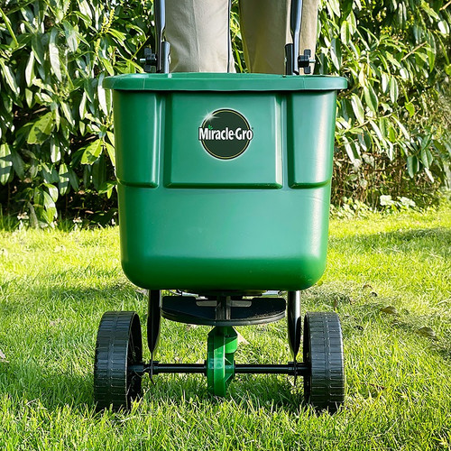 Green fertilizer spreader labeled "Miracle-Gro" on a lawn. Two wheels, standing upright, with blurred greenery in the background, conveying outdoor use.