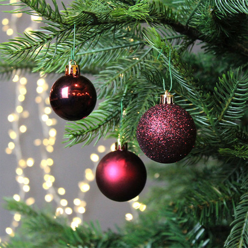 Burgundy Christmas baubles hanging on a fir tree, featuring glossy, matte and glitter finishes with warm white fairy lights in the background.