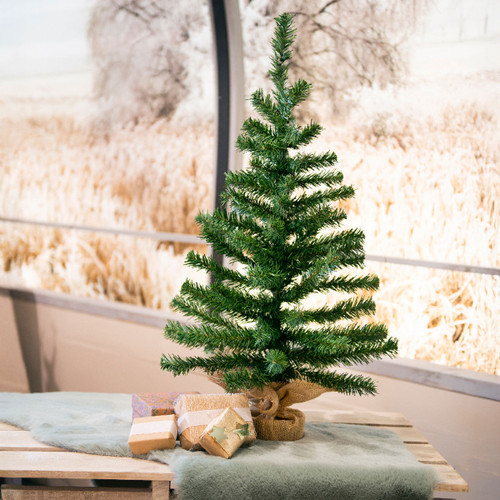 Small artificial tabletop Christmas tree displayed beside wrapped presents, featuring realistic green pine branches and a rustic burlap-wrapped base.