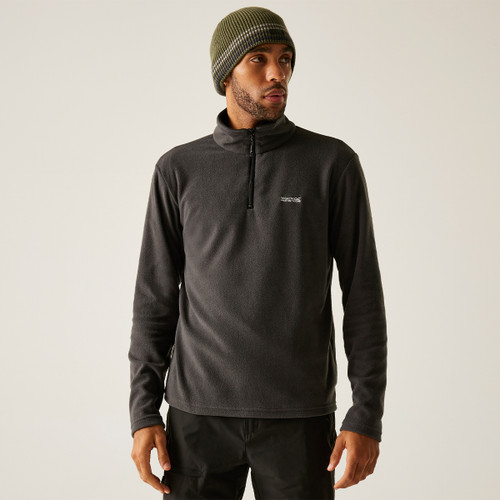 Man wearing a dark fleece pullover with a half-zip and a logo on the chest, paired with black pants and a green-striped beanie, against a beige background.