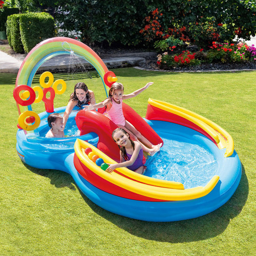 Children playing in a rainbow-themed inflatable water play centre with slide, splash pool and ring toss games on a lawn