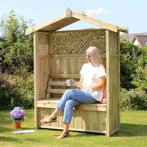 Wooden garden arbour with a pitched roof, lattice panels and bench seating with built-in storage, shown in a garden with a woman sitting and holding a mug.