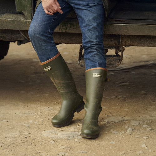 Man wearing green Barbour wellington boots standing on muddy ground beside a vehicle, shown with rolled blue jeans.