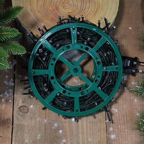 Christmas LED string lights wound on a green storage reel, displayed on a rustic wooden surface with artificial snow and festive greenery, ready for seasonal decorating and easy storage.