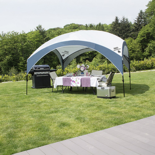 A spacious outdoor dining area under a canopy, featuring a table set with floral decor, chairs, a barbecue grill, and a cooler on green grass.
