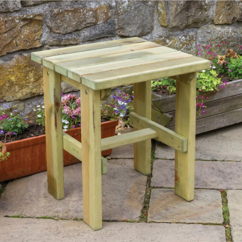 Wooden garden side table with a slatted top and sturdy legs, positioned on a paved patio beside a stone wall and flower planters.