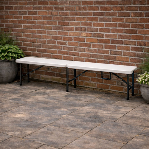 An elongated white folding bench with black metal legs, ideal for gatherings or events, against a brick wall.