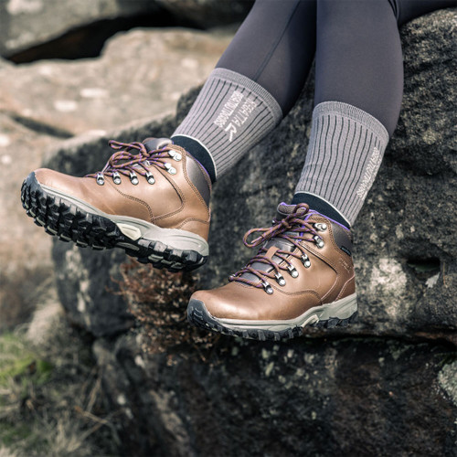 Brown leather hiking boots worn with grey walking socks and black leggings while sitting on rugged rocks outdoors.