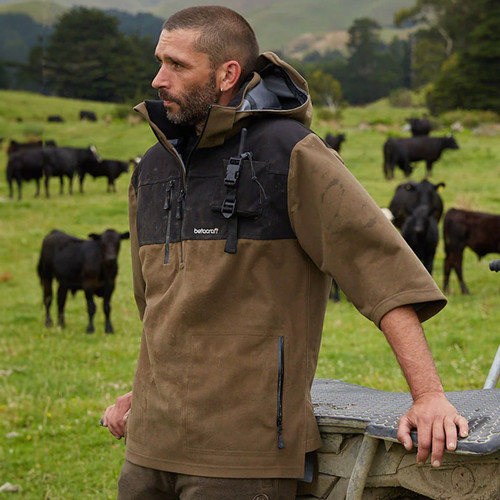 A man in a brown and black jacket stands in a green field, leaning on a fence, with several black cows grazing in the background on a cloudy day.
