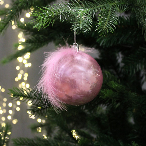 A pink bauble with a feather at the top, hanging from a Christmas tree branch, with gold string lights in the background.