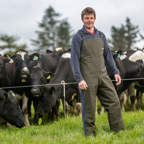A farmer in overalls stands on lush green grass, smiling in front of a herd of black and white cows. Overcast sky and trees in the background.