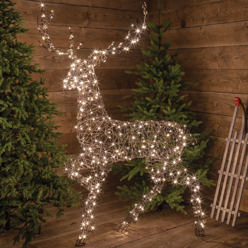 A wire framed wicker reindeer with white LEDs. The reindeer is in front of two Christmas trees