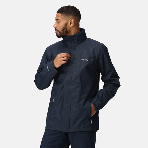 A man stands in a navy waterproof jacket with a high collar, looking to his left. His right hand holds the collar. The background is plain white.