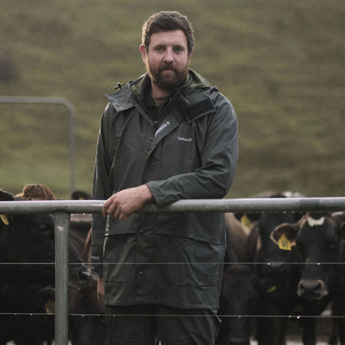 A man in a dark raincoat stands by a fence in a rainy field, with cows behind him.