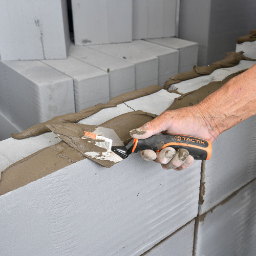 A hand uses a Tactix tool to spread mortar on a partially built wall of light grey concrete blocks. The scene conveys precision and craftsmanship.