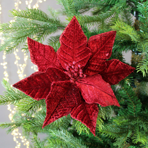 A vibrant red poinsettia ornament with textured petals is attached to a lush green Christmas tree branch, creating a festive and warm holiday atmosphere.