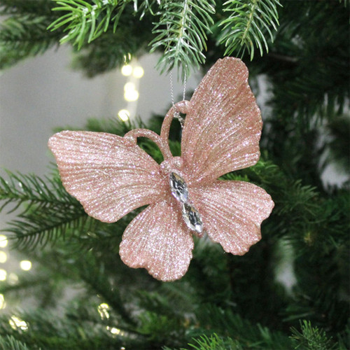 A blush pink glitter butterfly with a diamante body, attached to a Christmas tree branch with gold string lights in the background.