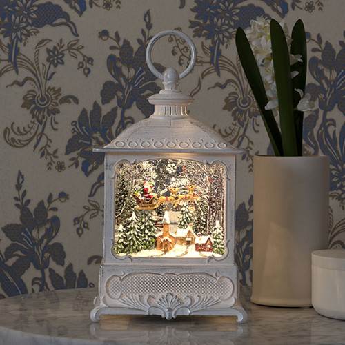 Decorative white Christmas lantern with illuminated winter woodland scene, featuring snow-covered trees, a deer, and warm LED lighting displayed on a side table.