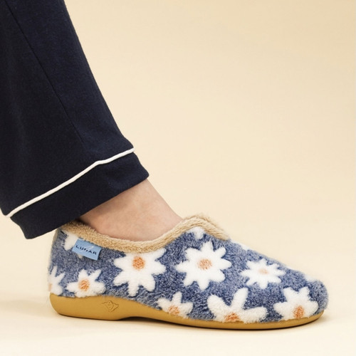 Side view of a foot wearing a cosy slipper with a blue fuzzy fabric covered in white and orange daisy patterns, trimmed with beige faux fur, and set against a beige background. The wearer is dressed in navy pyjamas with white piping.
