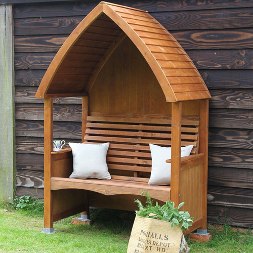 Stained beech wood cottage arbour bench with curved slatted roof and slatted back, styled with cushions beside a dark timber wall.