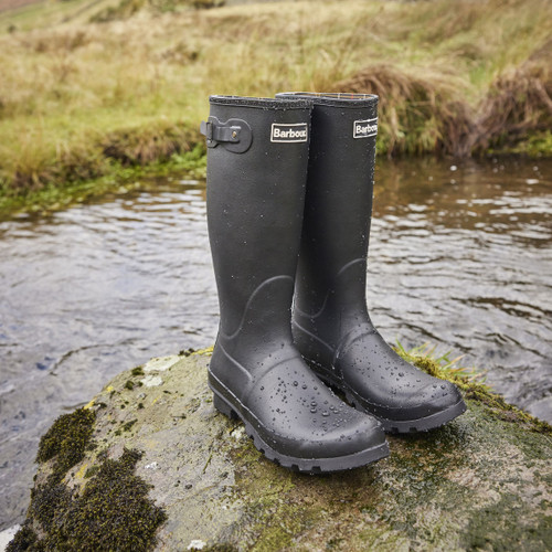 Pair of black Barbour wellington boots standing on a mossy rock beside a stream, water droplets visible on the rubber.