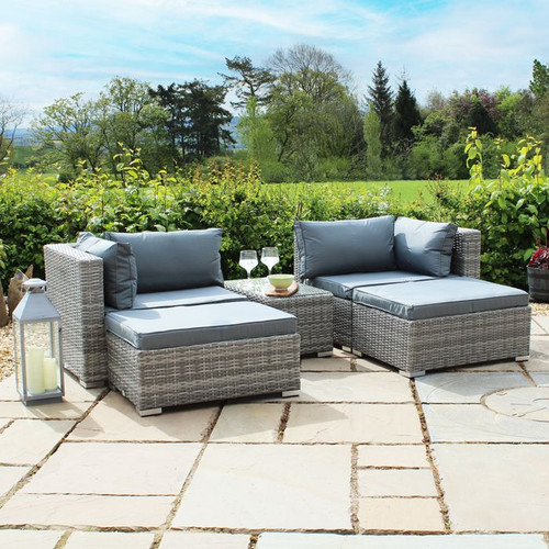 Two cushioned, grey wicker patio chairs with a small matching table on a stone patio, surrounded by lush greenery and under a bright blue sky.