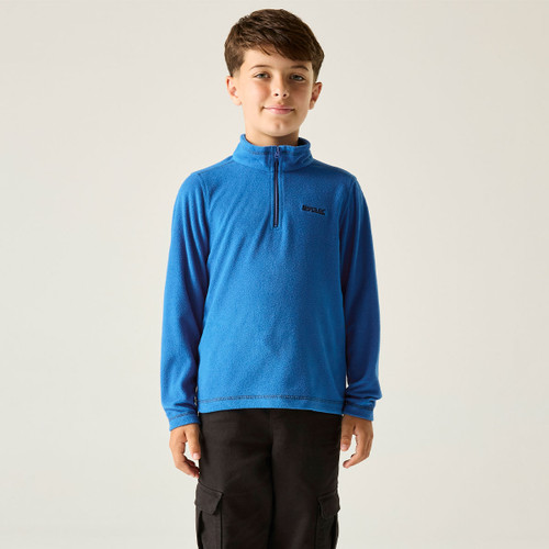 Young boy wearing a bright blue long-sleeve pullover with a quarter-zip and black cargo pants. He stands against a plain, light background, smiling softly.