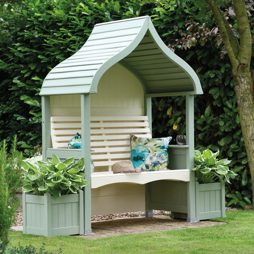 Sage and cream painted garden arbour with curved roof, slatted backrest and built-in planter boxes, styled with cushions in a leafy garden.