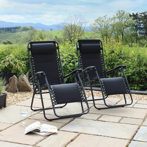 Two black reclining chairs sit on a stone patio, surrounded by greenery and rolling hills under a cloudy sky. A book lies open nearby.