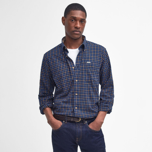 Man posing in a casual outfit: a blue and orange plaid button-up shirt over a white T-shirt, with dark jeans. Hands in pockets, neutral expression.