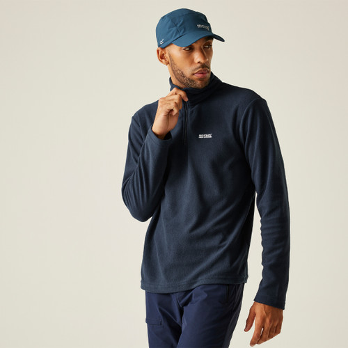Man in navy fleece pullover and matching cap, with relaxed stance against plain background. He gazes to the side, conveying a casual, thoughtful tone.