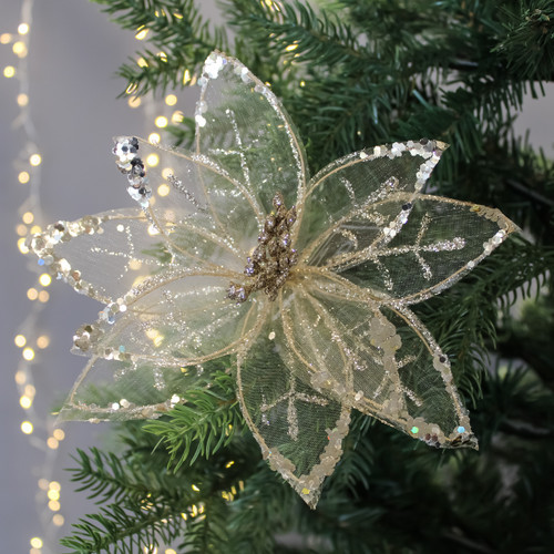Sparkling gold poinsettia ornament with translucent petals and sequins, set against lush green pine needles and twinkling fairy lights, evokes festive elegance.