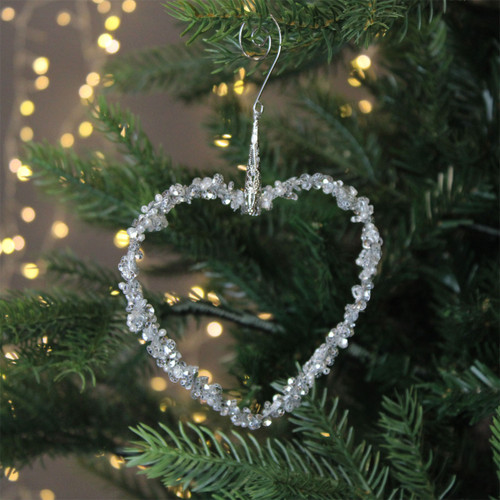 A silver beaded heart decoration hanging from a Christmas tree, with gold string lights in the background.