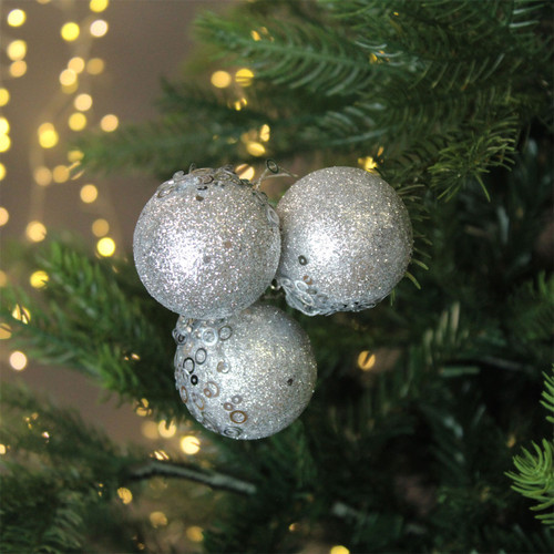 A cluster of three silver glitter balls hanging from a Christmas tree, with gold string lights in the background.