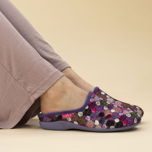 A person wearing purple Lunar mule slippers with a colourful patchwork knit design featuring circular polka dots in shades of pink, purple, green, and white. The slippers have matching purple soles and trim, and are paired with mauve ribbed loungewear trousers.