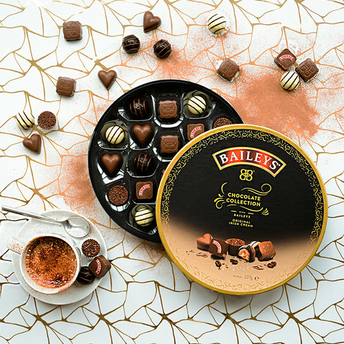 Opened tin of Baileys Chocolate Collection with assorted milk, dark and white chocolates displayed on a patterned surface with cocoa powder.