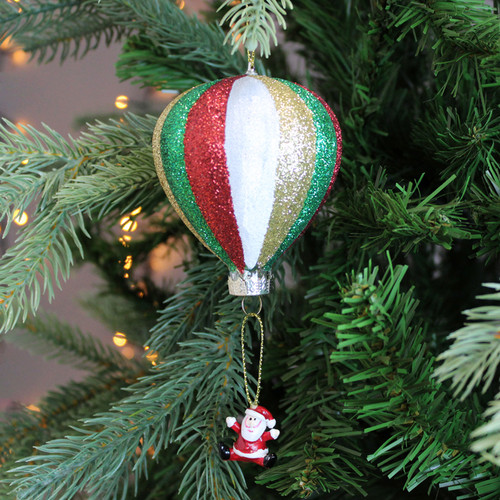 A hot air balloon decoration with glitter segments, carrying Santa through the air. The decoration is hanging from a Christmas tree branch, with gold string lights in the background.