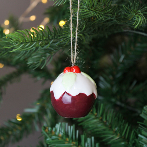 A ceramic Christmas pudding decoration hanging from a Christmas tree, with gold string lights in the background.