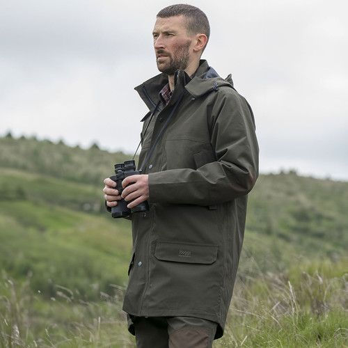 Man wearing a dark olive green waterproof outdoor jacket while holding binoculars in a countryside setting.