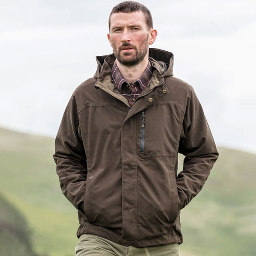 Man wearing a dark olive hooded jacket, zipped up with his hands in the side pockets, standing against a backdrop of green hills.