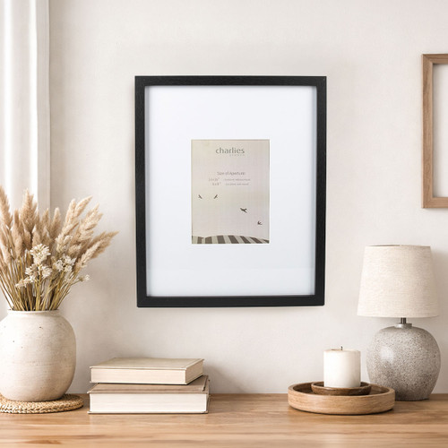 Black photo frame with white mount displayed on a neutral wall above a wooden table, styled with books, a ceramic vase of dried grasses, a candle and a table lamp in a calm home interior.