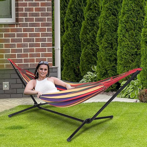 Person relaxing in a colourful striped hammock with a black metal stand on a garden lawn beside a brick house.