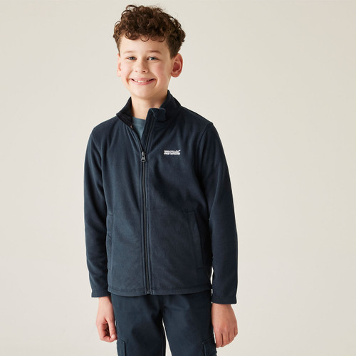 A smiling boy in a navy fleece jacket and matching pants stands against a plain background, conveying a cheerful and relaxed mood.