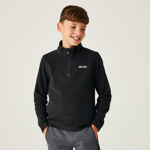 A smiling boy in a black half-zip pullover and grey pants stands with hands in pockets against a plain background, exuding a casual and happy vibe.