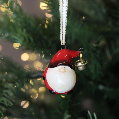 A round Santa decoration with a white beard and a red hat with a bell, hanging from a Christmas tree with gold string lights in the background.