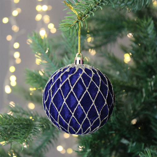 A deep blue Christmas ornament with a silver diamond pattern hangs on a pine tree, surrounded by soft, glowing lights, creating a festive atmosphere.