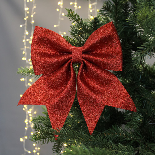 A large, glittering red bow decorates a Christmas tree, set against softly glowing string lights. The scene conveys a festive, warm holiday spirit.