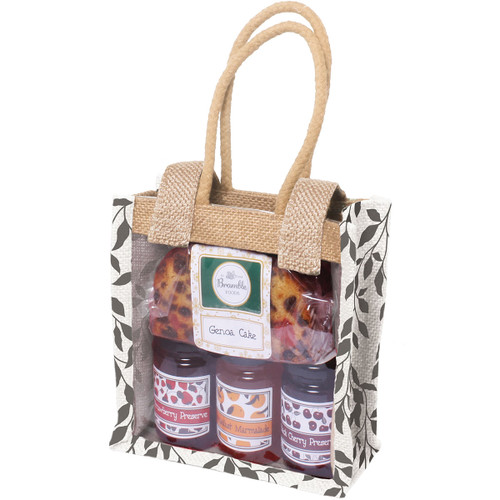 Gift bag with woven handles containing a fruit cake and three jars of preserves, visible through a transparent front. The bag has a black leaf pattern.