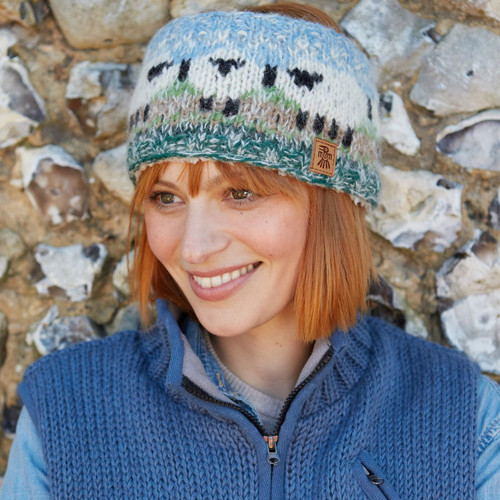 Woman smiling in front of a stone wall, wearing a colourful knitted headband with sheep design and a blue vest. The scene conveys a cheerful, cosy vibe.